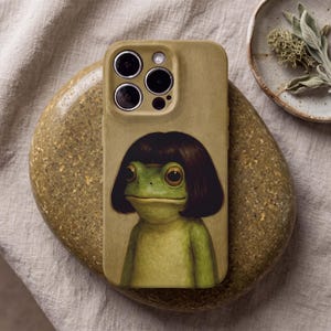May include: A tan smartphone case featuring a printed illustration of a green frog with large eyes and a dark brown bob haircut. The case has a cutout for a triple-lens camera system and a flash, maintaining a smooth, matte-textured finish throughout.