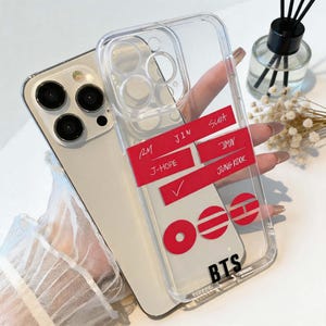 Könnte beinhalten: Transparente Handyhülle mit rotem Design mit den Namen RM, J-HOPE, Jin, Suga, Jimin und Jung Kook. Die Hülle hat auch die Buchstaben BTS und ein Häkchen.