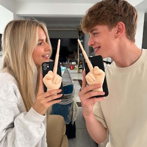Puede incluir: Una funda de smartphone negra que presenta una escultura de mano tridimensional de tono claro con el dedo corazón extendido. La mano está colocada verticalmente en la parte posterior de la funda, sirviendo como elemento decorativo.
