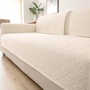 Könnte beinhalten: Ein cremefarbener Sofabezug mit einem strukturierten Webmuster aus sich wiederholenden diagonalen Linien. Der Bezug liegt über den Sitz- und Rückenkissen des Sofas und sorgt für ein einheitliches, helles Erscheinungsbild auf der Möbeloberfläche.