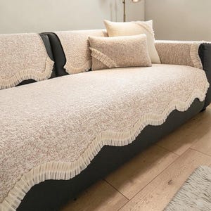 May include: A beige and white textured sofa cover set featuring a scalloped edge with pleated ruffles. The set includes a large main cover draped over the seat and back, along with two rectangular throw pillows with matching ruffled trim details.