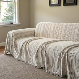 May include: A cream-coloured sofa with a matching throw blanket. The blanket has a chevron pattern in a darker neutral colour. The sofa is in front of a window and a neutral-coloured wall. The throw blanket has a fringed edge.