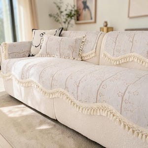 May include: A cream sofa with a floral patterned cover and matching cushions. The cover has a scalloped edge with cream-coloured fringe. Two cushions are on the sofa, one with a black and white design and the other with a floral pattern.