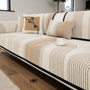 Puede incluir: Un sofá color crema con un patrón repetitivo de líneas curvas marrones. El sofá tiene varias almohadas decorativas en colores y patrones complementarios. Una taza y un platillo blancos están sobre el sofá.