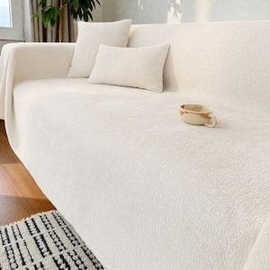 Könnte beinhalten: Ein cremefarbenes Sofa mit passenden Kissen und einem strukturierten Überwurf. Eine kleine, dekorative Tasse mit braunem und beigem Design steht auf dem Sofa. Das Sofa steht auf einem Holzboden, mit einem gemusterten Teppich.