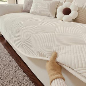 Puede incluir: Sofá color crema con un patrón geométrico acolchado. El sofá tiene cojines a juego y un cojín decorativo en forma de flor. Una mano con un guante beige ajusta la funda del sofá.