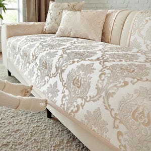 Può includere: Copridivano beige con un motivo damascato dettagliato nei toni crema e oro. La copertura è posta su un divano bianco, con cuscini abbinati. Il design generale è elegante e classico, adatto all'arredamento del soggiorno.