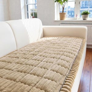 Könnte beinhalten: Eine beige, strukturierte Sofabezug mit einem Steppmuster, platziert auf einem weißen Sofa. Der Bezug hat einen gerippten Rand und dient zum Schutz des Sofas. Der Raum wird durch natürliches Licht von einem Fenster erhellt.