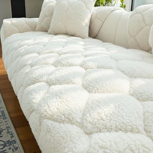 May include: A close-up of a white, textured sofa cover and matching pillows. The cover has a quilted pattern, and the fabric appears soft and plush. The pillows have a similar design, creating a cohesive look. The overall aesthetic is cozy and inviting.