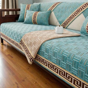 Könnte beinhalten: Ein türkisfarbenes Sofa mit passenden Kissen und einem gemusterten Design. Das Sofa hat einen braunen Rand mit einem geometrischen Muster. Eine beige Decke und eine weiße Teekanne befinden sich auf dem Sofa.