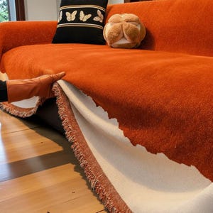 May include: An orange sofa blanket with a fringed edge is being lifted to reveal a cream-coloured underside. A black cushion with gold butterfly embroidery and a tan decorative cushion are on the sofa.