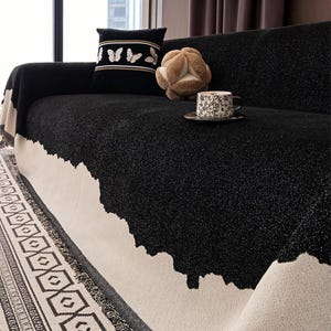 May include: A black sofa is draped with a textured throw blanket with a white border. A decorative pillow with butterfly embroidery and a plush, spherical object rest on the sofa. A patterned teacup sits on a saucer.