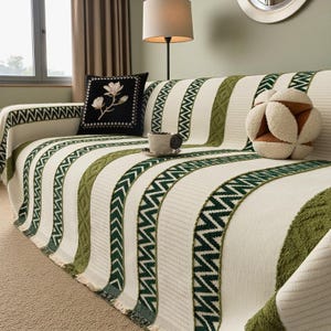 Può includere: Una fodera per divano color crema con strisce verdi e motivi a zig-zag. La fodera, con un bordo a frange, è drappeggiata su un divano. Un cuscino nero con un motivo floreale e un cuscino decorativo a sfera sono sul divano.