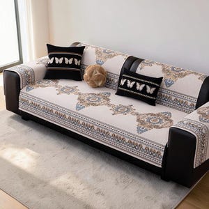 Könnte beinhalten: Ein dunkelbraunes Sofa mit einem hellbeigen Stoffbezug, der ein sich wiederholendes Muster aus blauen und goldenen Medaillons aufweist. Das Sofa enthält zwei schwarze quadratische Kissen mit weißen Schmetterlingsmotiven und ein kleines beiges Kugel-Kissen.