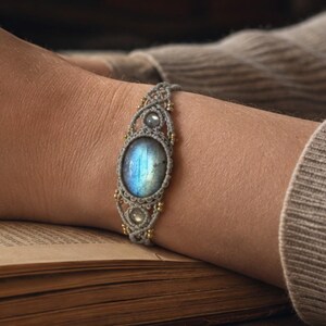 May include: A macrame bracelet featuring a large, oval-shaped labradorite gemstone with a blue iridescent sheen, accented by smaller round stones and gold-toned beads, woven with grey cord in an intricate, knotted pattern around the wrist.