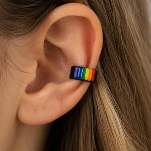 May include: A small ear cuff featuring a vibrant rainbow-colored striped pattern. The accessory is designed with a black base and vertical bands of red, orange, yellow, green, blue, and violet colors, creating a distinct and colorful appearance.