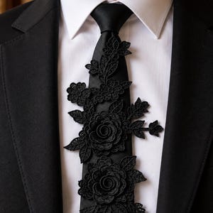 May include: A black necktie featuring intricate black lace floral appliqués layered over the fabric. The tie is worn with a crisp white collared shirt and a black suit jacket, creating a formal appearance with detailed embroidery textures.