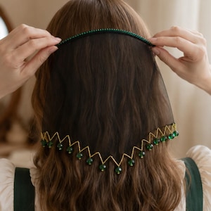 May include: A black sheer veil headband featuring a thin band adorned with small green beads. The lower edge of the veil is trimmed with a gold-toned zigzag pattern, with each point finished with a dangling dark green bead accent.