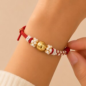 Könnte beinhalten: Rot-weiß geflochtenes Armband mit goldfarbenen Kugeln und Akzenten. Das Armband hat einen zentralen goldfarbenen Ball und kleinere weiße Perlen, wodurch ein dekoratives Handgelenkaccessoire entsteht.