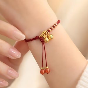 May include: Red braided bracelet with gold-colored accents and two small bells. The bracelet features a knot design and two dangling red beads. The bracelet is worn on a wrist.
