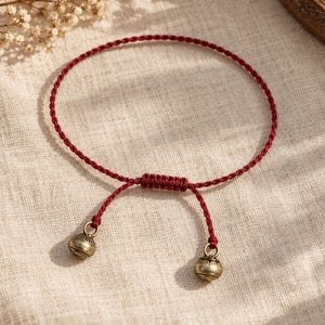 May include: A thin, braided dark red cord bracelet featuring an adjustable sliding knot closure. Two small, round, antique brass-toned metal bells are attached to the ends of the cords, hanging down from the central knot mechanism.