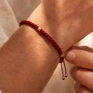 Op de afbeelding: Een rood gevlochten armband met goudkleurige kralen. De armband is vastgemaakt met een schuifknoopsluiting en heeft twee kleine gouden kralen aan de uiteinden. De armband wordt om de pols gedragen.
