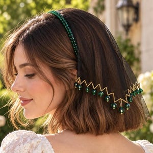 May include: A black tulle veil headband featuring a band adorned with small green beads. The lower edge of the black netting is trimmed with a gold-toned zigzag chain, accented by dangling dark green spherical beads along the entire border.