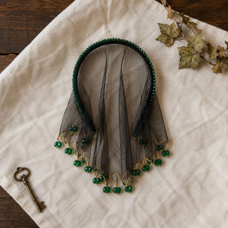 May include: A black headband featuring a sheer black tulle veil. The headband is adorned with small green beads along the frame, and the bottom edge of the veil is decorated with gold thread and dangling green faceted beads.