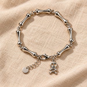 Könnte beinhalten: Ein silberfarbenes Kettenarmband mit einer Reihe polierter, knochenförmiger Glieder, die durch kleine Metallringe verbunden sind. Das Armband verfügt über einen Karabinerverschluss, eine verstellbare Verlängerungskette mit runder Plakette und einen Totenkopf-Anhänger.