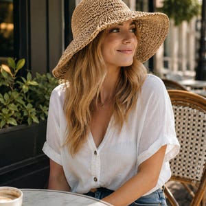 May include: A tan woven straw sun hat with a wide, flexible brim paired with a white short-sleeved button-down shirt featuring a V-neckline and cuffed sleeves, worn with blue denim jeans and a simple thin metallic necklace with a small circular pendant.