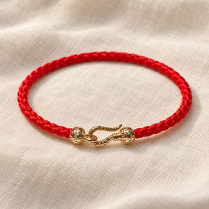 以下が含まれることがあります： 鮮やかな赤色の編み込みコードブレスレットで、ゴールドトーンの金属製フッククラスプが付いています。クラスプは2つのテクスチャ加工された球状のビーズで構成され、表面全体に細かい凹凸のある曲線的なフック型の留め具で接続されています。
