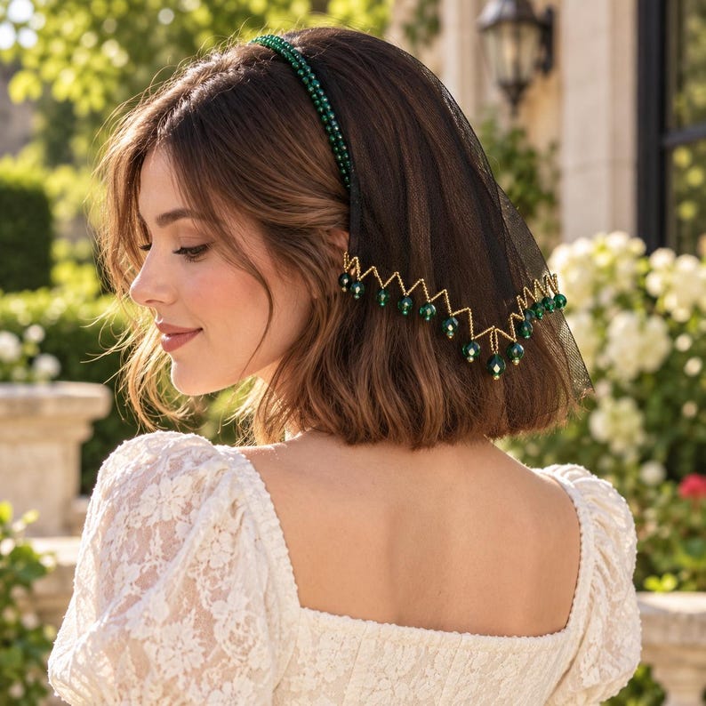 May include: A black tulle veil headband featuring a band adorned with small green beads. The lower edge of the black netting is trimmed with a gold-toned zigzag chain, accented by dangling dark green spherical beads along the entire border.