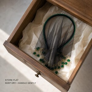 May include: A black headband featuring a sheer black veil and a border of small green beads. The lower edge of the veil is decorated with dangling green beads attached by thin gold-toned wire. Text reads: STORE FLAT, KEEP DRY, HANDLE GENTLY.