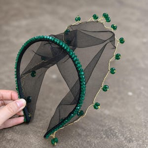 May include: A black headband featuring a band covered in small green faceted beads and a layer of sheer black tulle. The tulle is edged with a thin gold wire adorned with spaced green beads, creating a decorative, layered, and textured appearance.