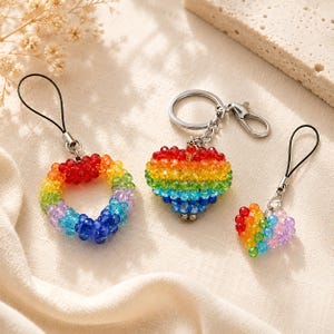 May include: Three rainbow-coloured beaded keyrings. One is a circle, another a heart, and the third is a smaller heart. Each keyring has a silver-coloured clasp or loop for attachment.