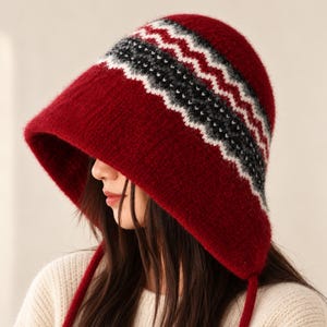 May include: A deep red knit hat with a fuzzy texture and ear flaps. The hat features a prominent horizontal band across the crown decorated with a white, gray, and black zigzag pattern. Two long red knit ties hang down from the ear flaps.