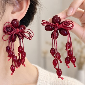 May include: A pair of deep red earrings featuring a central cluster of smooth, oval-shaped beads arranged like a flower. A decorative red cord is tied into a bow at the center, with multiple long, dangling strands ending in individual red beads.