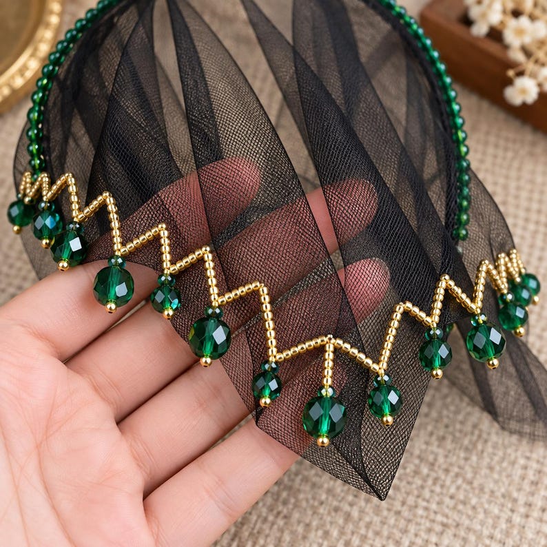 May include: A headband featuring a black tulle veil, adorned with a zigzag pattern of small gold beads and dangling faceted emerald green crystal beads. The headband frame is lined with a row of tiny green beads along the outer edge.
