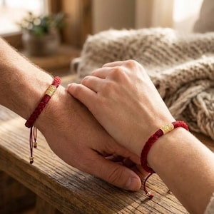 May include: Two red braided bracelets with gold-colored cylindrical accents are shown on wrists. The bracelets are tied with adjustable cords. The image shows two people holding hands, with the bracelets prominently displayed on their wrists.