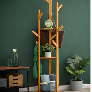 Puede incluir: Un perchero de madera con diseño de ramas de árbol. Cuenta con estantes, ganchos y una sección superior para colgar objetos. Se exhiben un reloj verde, un bolso tote marrón y una planta en maceta. También se ven una bufanda verde y toallas azules.