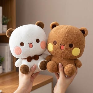 May include: Two plush bear toys. One is white with brown accents and a bow tie, the other is brown with yellow cheek circles. Both have smiling faces with pink cheeks and small tongues.