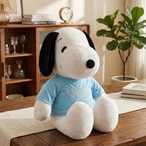 Può includere: Un peluche di Snoopy, principalmente bianco con orecchie nere e naso nero, indossa una maglietta azzurra con la scritta "SMILED" ricamata. La bambola è seduta su un tavolo di legno.