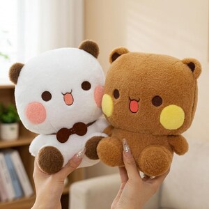 May include: Two plush bear toys. One is white with brown ears, a brown bow tie, and pink cheeks. The other is brown with yellow cheeks. Both have black eyes and open mouths with red tongues. The toys are held up by hands.