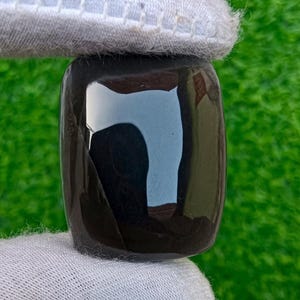 Op de afbeelding: Een gepolijste, rechthoekige zwarte edelsteen met afgeronde randen wordt vastgehouden door een gehandschoende hand. Het glanzende oppervlak reflecteert licht. De achtergrond is heldergroen. De edelsteen is waarschijnlijk voor sieraden of decoratieve doeleinden.