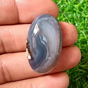 Może przedstawiać: Owalny kamień szlachetny o wypolerowanej powierzchni. Kamień ma wirujące wzory w odcieniach szarości, bieli i błękitu. Kamień szlachetny jest trzymany w dłoni, z widocznym zielonym tłem.