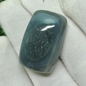 May include: A polished, rectangular gemstone with a translucent, teal-blue hue. The stone features a unique, speckled pattern within its depths, creating a captivating visual texture. The gemstone is held against a white fabric background.