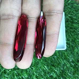 Pode incluir: Duas pedras preciosas vermelho-escuras alongadas com corte marquise e superfícies facetadas que refletem a luz. As pedras são mantidas verticalmente, exibindo um tom carmesim vibrante e um acabamento polido e transparente.