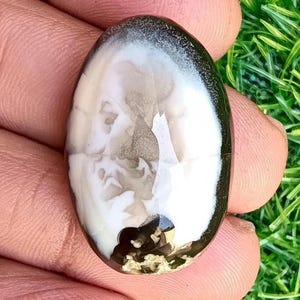 May include: An oval-shaped gemstone with a polished surface. The stone features a striking contrast of black, white, and gray hues. The design has a marbled appearance, with swirling patterns and a dark border. The gemstone is held in a hand.