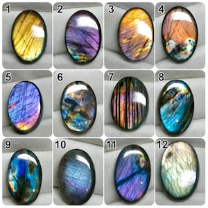 May include: A collection of twelve oval-shaped labradorite gemstones, each featuring unique iridescent flashes of color including gold, purple, blue, and orange. The stones are arranged in a grid, displaying natural surface textures and light reflections.