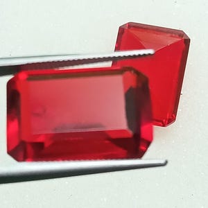 Op de afbeelding: Twee gefacetteerde, rechthoekige, rode edelstenen. Eén wordt vastgehouden door een pincet, de andere is erachter geplaatst. De edelstenen hebben een hoogglans afwerking en zijn geplaatst tegen een witte achtergrond. De edelstenen zijn een levendig, transparant rood.